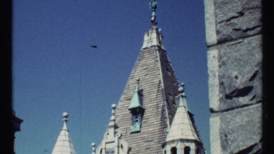 1967 8mm frame of a steep church roof with finial details, small turret forms, and a rough stone wall at the right edge.