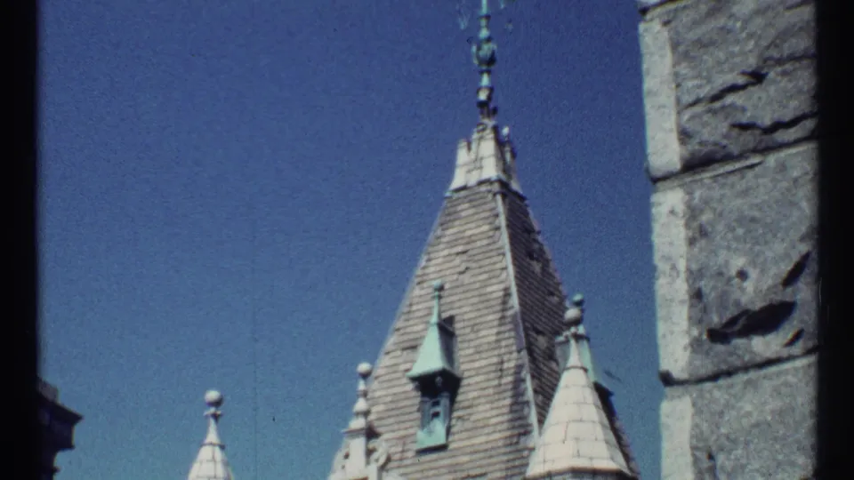 1967 8mm frame showing the upper portion of a pointed steeple, decorative spires, roof shingles, and a vertical stone wall edge.