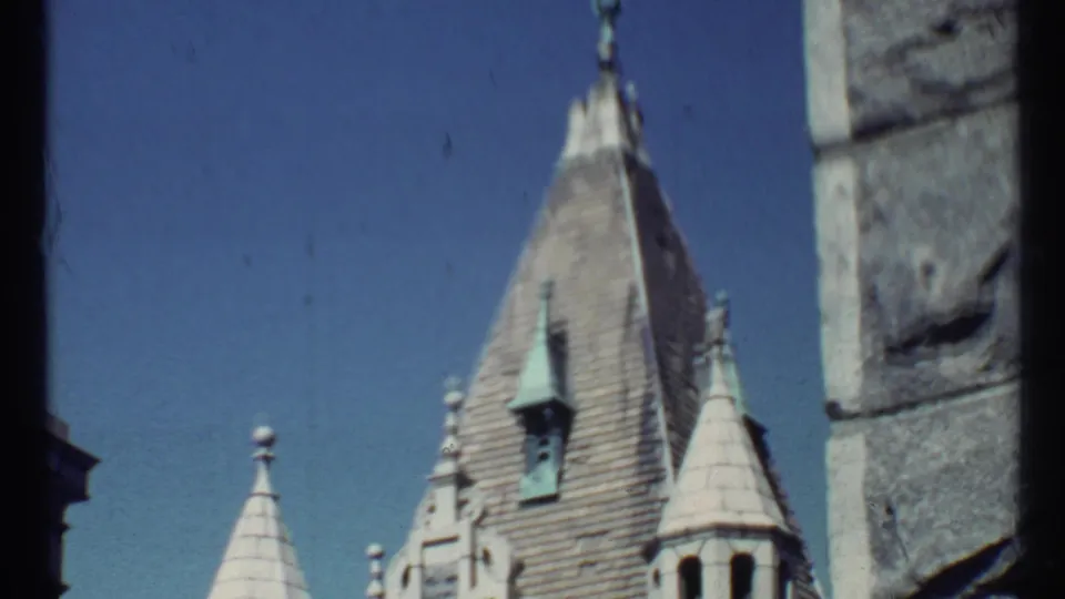 1967 8mm frame with a softer view of the church steeple, pale turret roof, blue sky, and rough stone blocks along the right side.