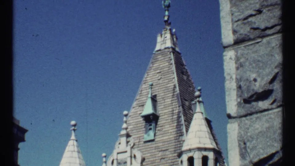 1967 Toronto home-movie frame showing steep Gothic-style roof spires, dormers, and a close stone wall against a clear sky.
