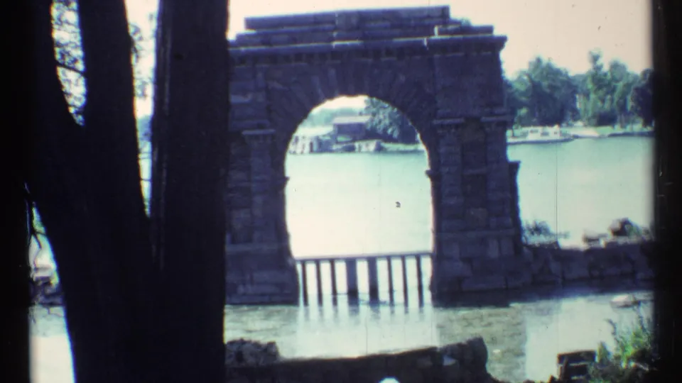 1967 Toronto home-movie still showing a stone arch and lakeside water, with trees and small boats on the far side.