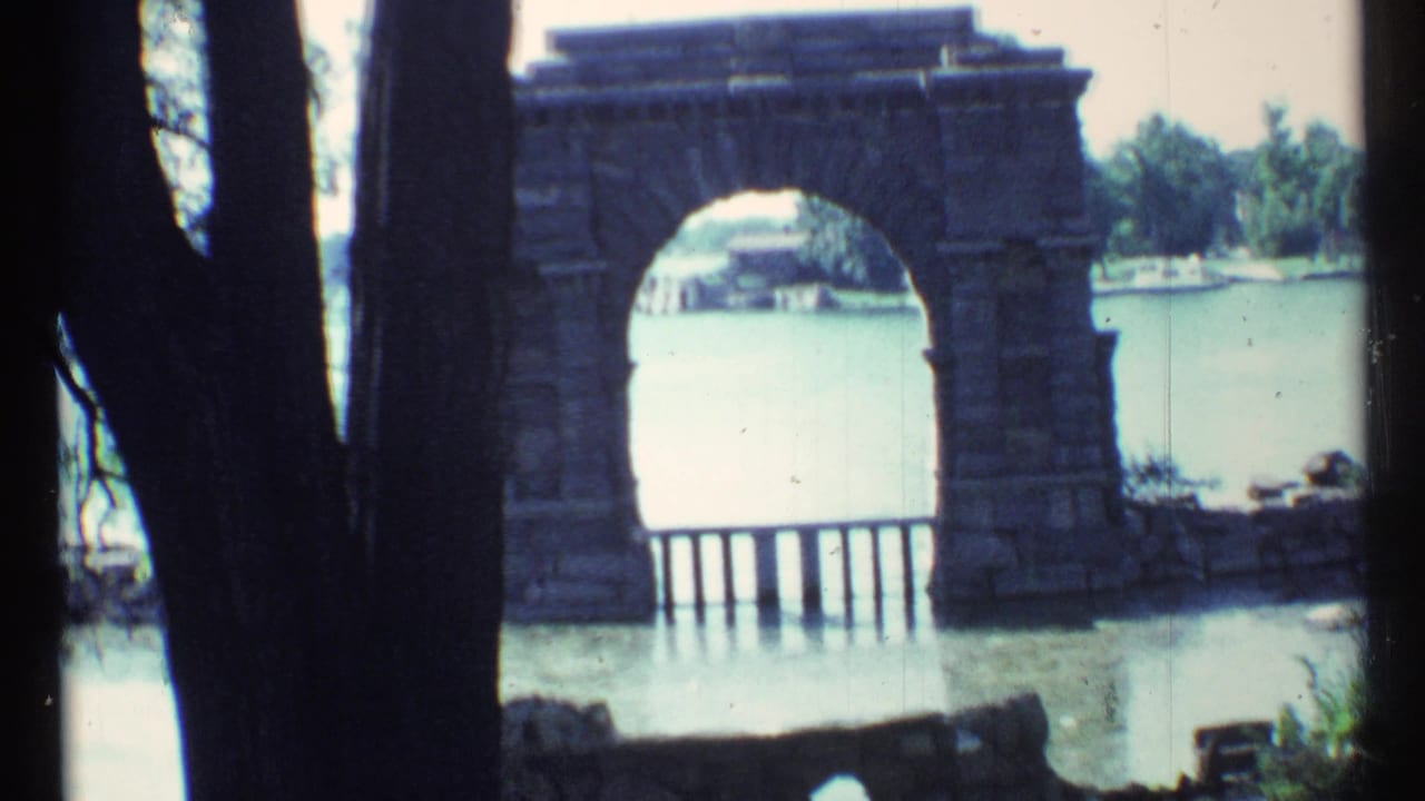 Stone archway beside water framed by dark tree trunks in bright daylight.