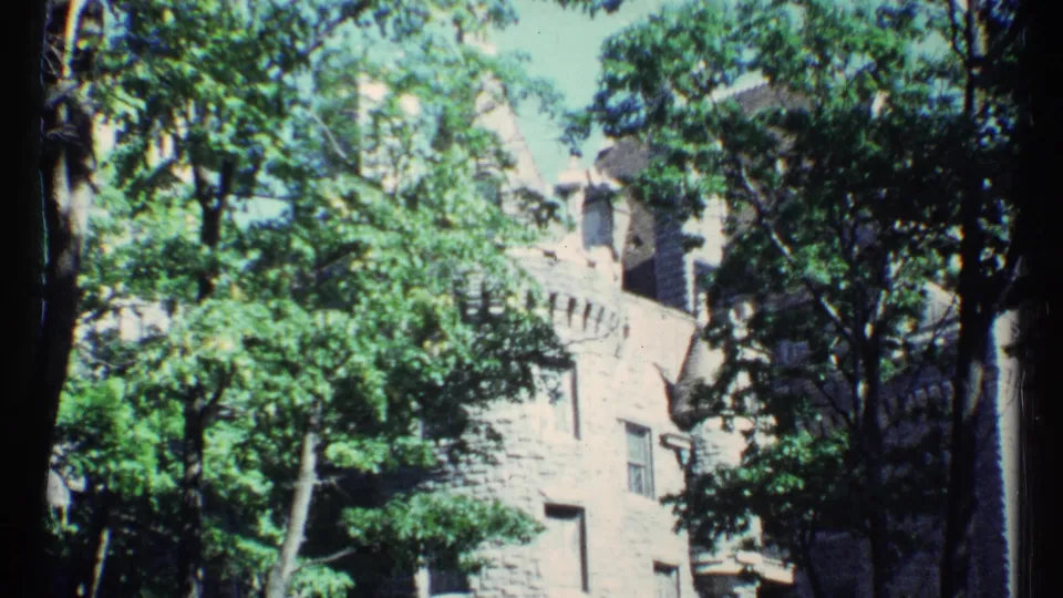 1967 Toronto footage frame showing a stone building with windows and roof details behind leafy trees in bright daylight.