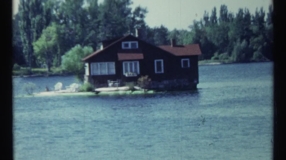 1967 Toronto home-movie view of a small lakeside cabin with red roof, white trim, chairs, and surrounding trees.