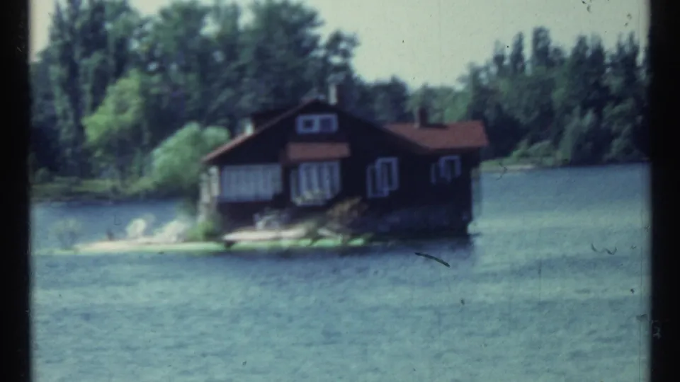 1967 Toronto frame showing the island cabin from across the water, with shoreline trees and soft 8mm focus.
