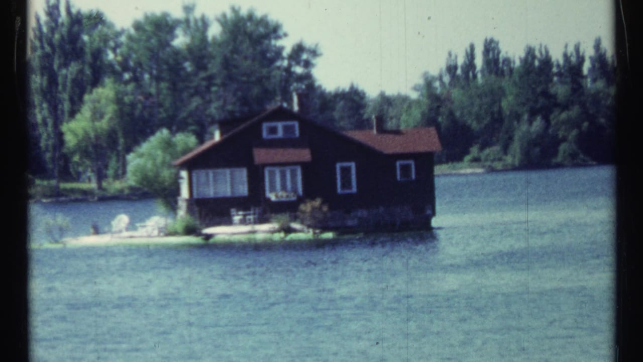 Dark wooden cabin on a small island beside calm lake water and trees.