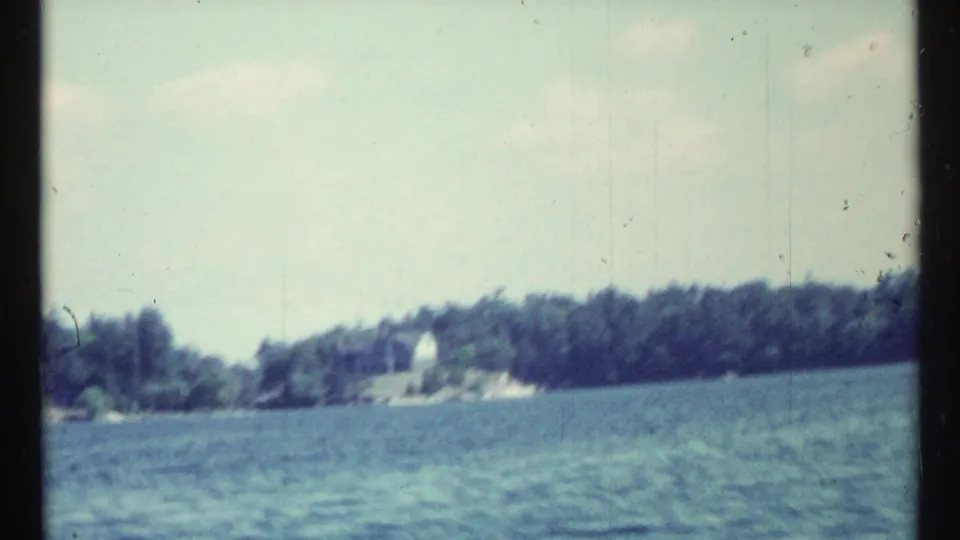 Soft 8mm frame showing open lake water, a distant tree-lined shore, and a small pale building or structure.