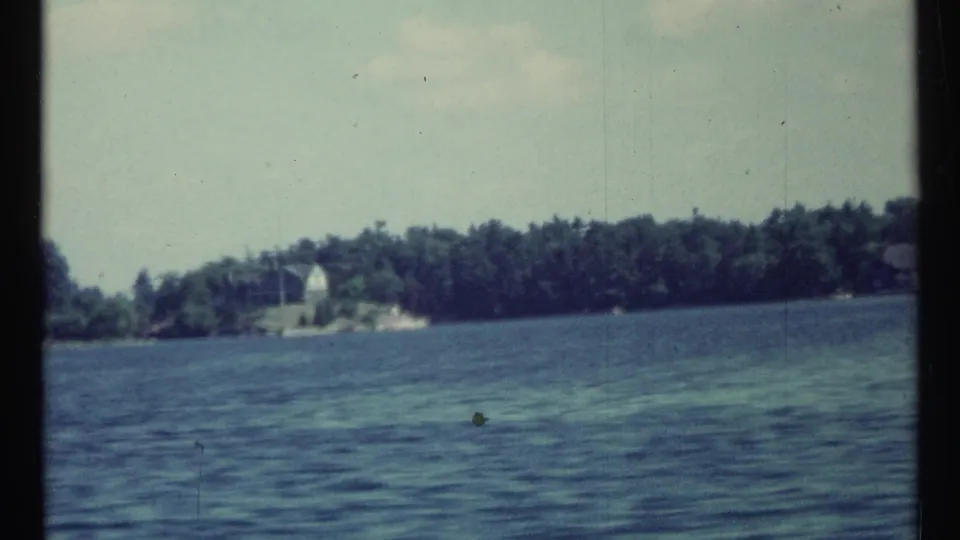 Wide 1967 8mm lake view near Toronto with rippled water, distant trees, sky, and a small pale shoreline structure.