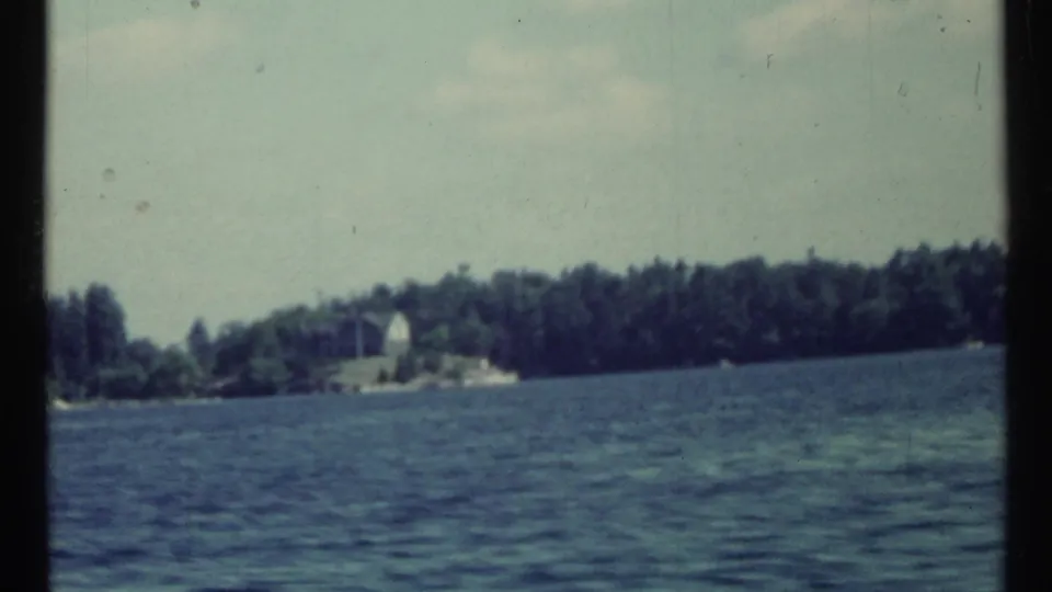 1967 8mm home-movie frame showing blue water, a wooded shoreline, and a small pale distant structure near Toronto, Canada.