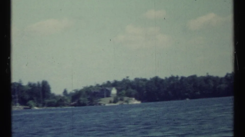1967 8mm frame with handheld motion blur, showing rippled water, a far wooded shore, and a small pale building-like shape.