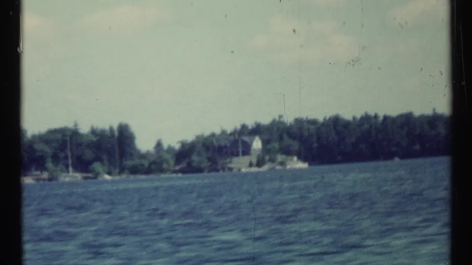 Final 8mm frame of the lakeside passage, with deep blue water, wooded shoreline, and a small white distant structure in view.