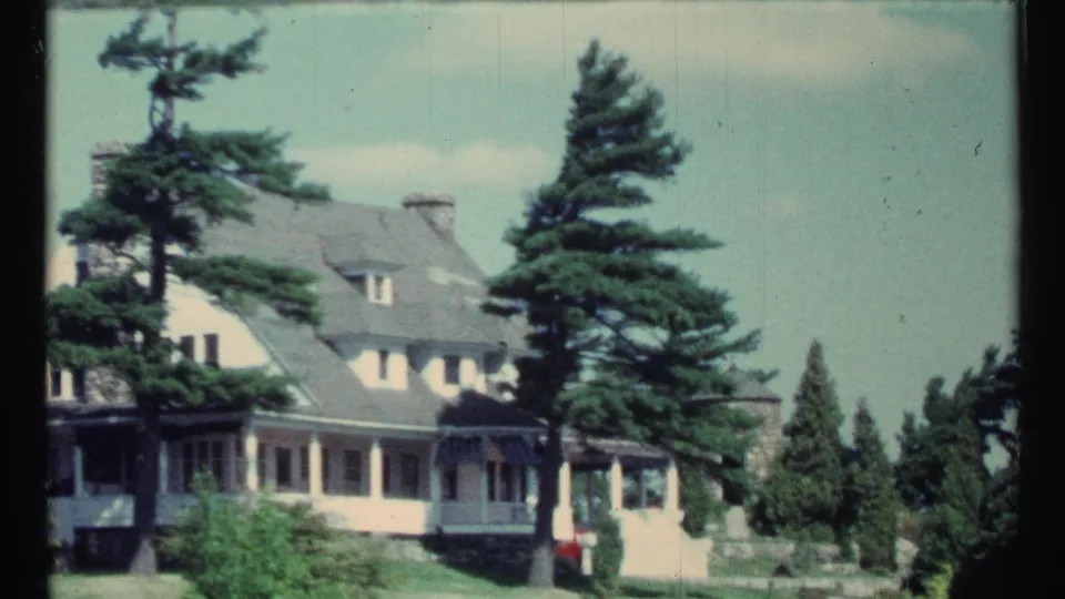 Closer 1967 8mm view of a white house with a porch and evergreen trees in a Toronto-area lakeside setting.