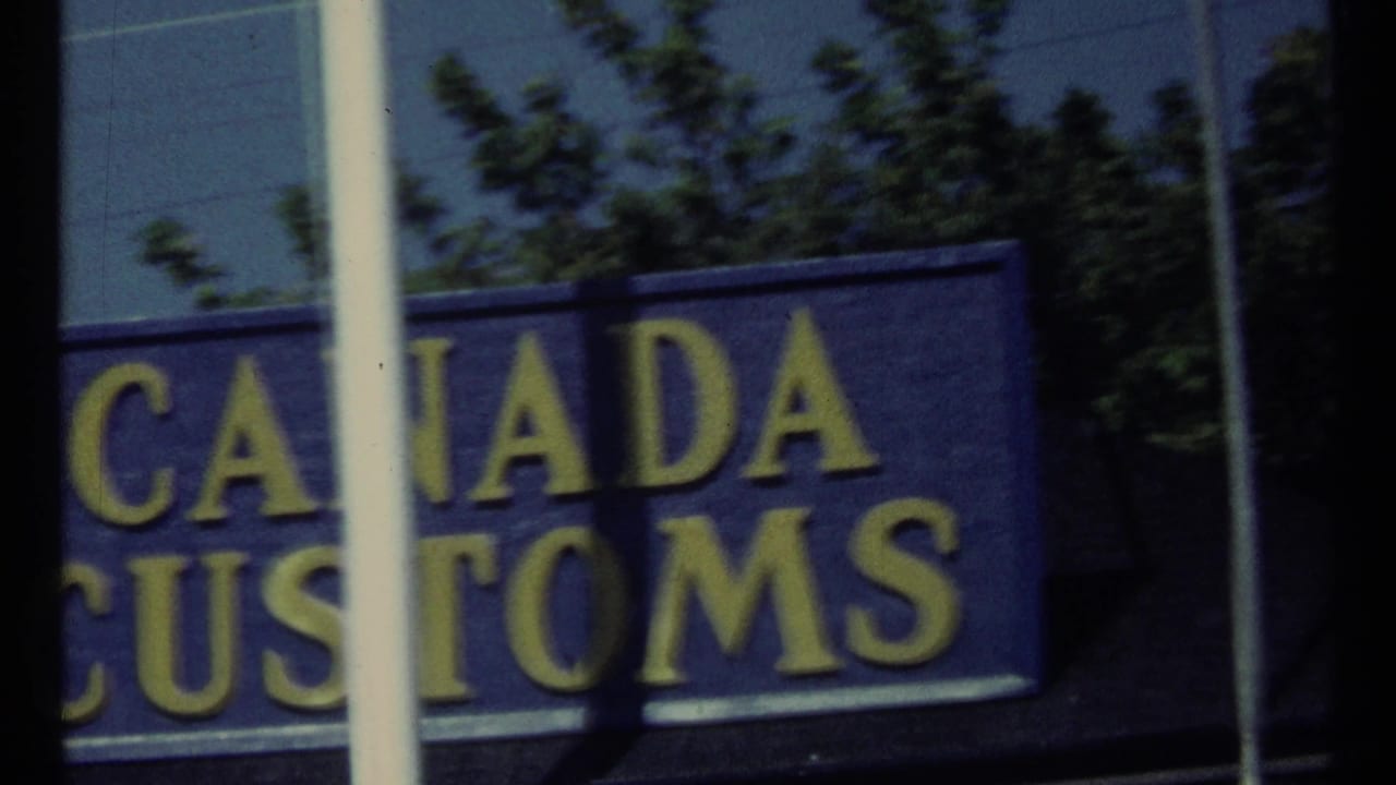 1967 Toronto home-movie frame showing part of a blue Canada Customs sign with yellow lettering through reflective glass.