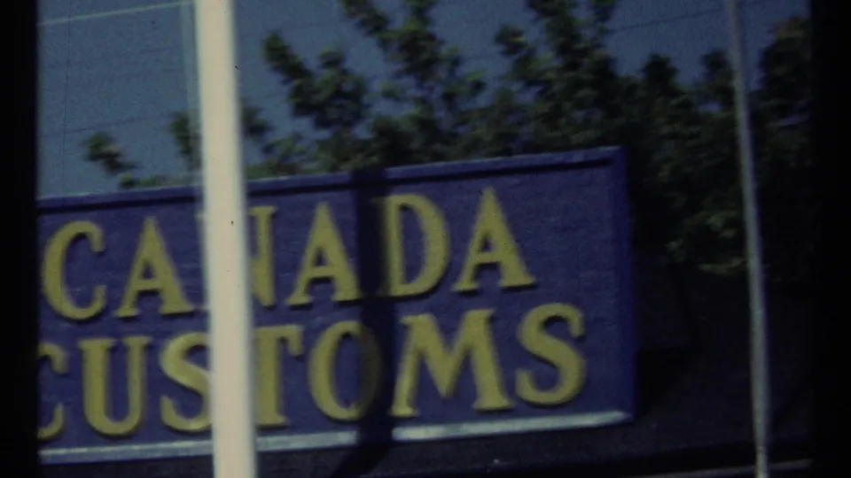 1967 Toronto home-movie frame showing part of a blue Canada Customs sign with yellow lettering through reflective glass.