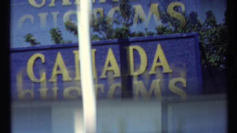 1967 Toronto customs sign viewed through reflective glass, with vertical bars, tree reflections, and soft 8mm blur.