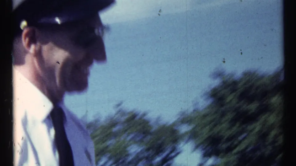 A uniformed adult man appears in profile in a soft, handheld 1967 Toronto-area home-movie frame with trees in the background.