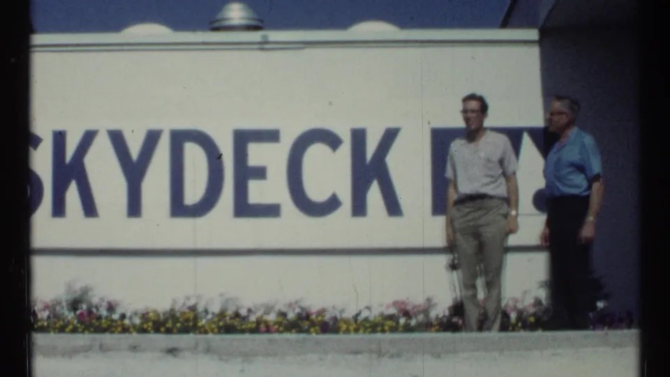 Two adult men stand in front of a white building with partial Skydeck text, flowers, and 1960s casual clothing visible.