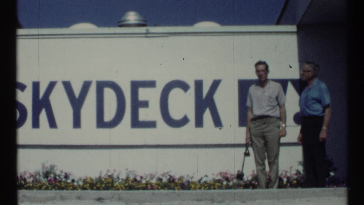 In Toronto in 1967, two adult men stand near a large Skydeck sign, with a flower bed and blue sky visible in 8mm footage.