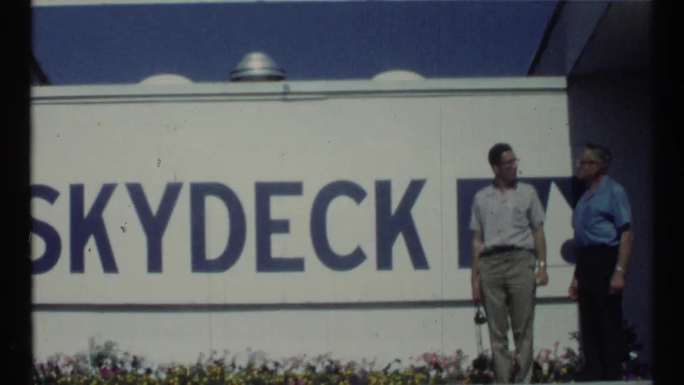 Two adult men appear to converse beside the blue-and-white Skydeck sign in 1967 Toronto home-movie footage.