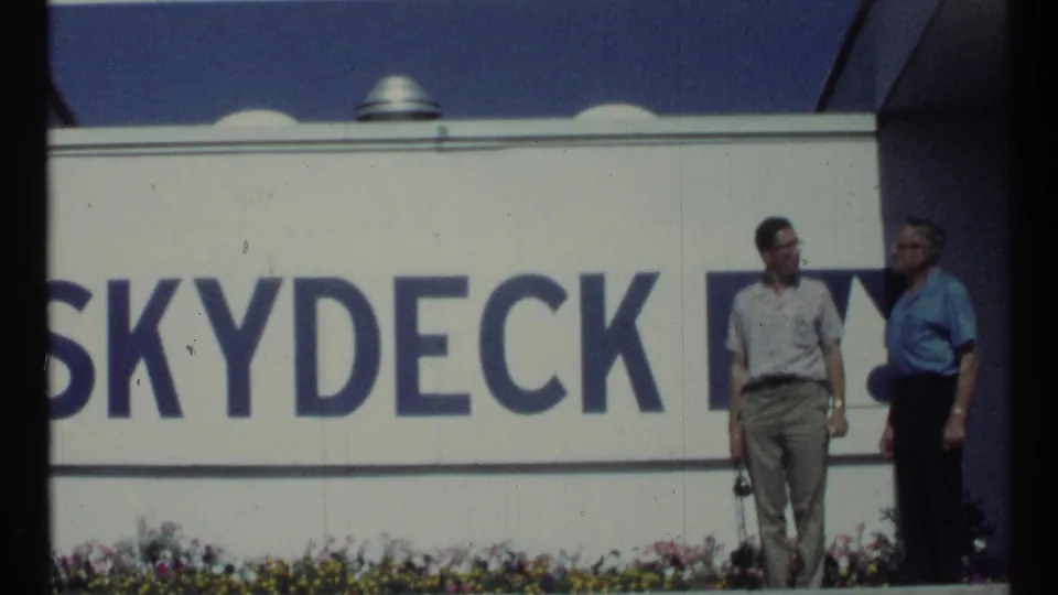 A visitor holding a small camera or strap stands with another man beside the Skydeck sign in Toronto, based on supplied 1967 metadata.