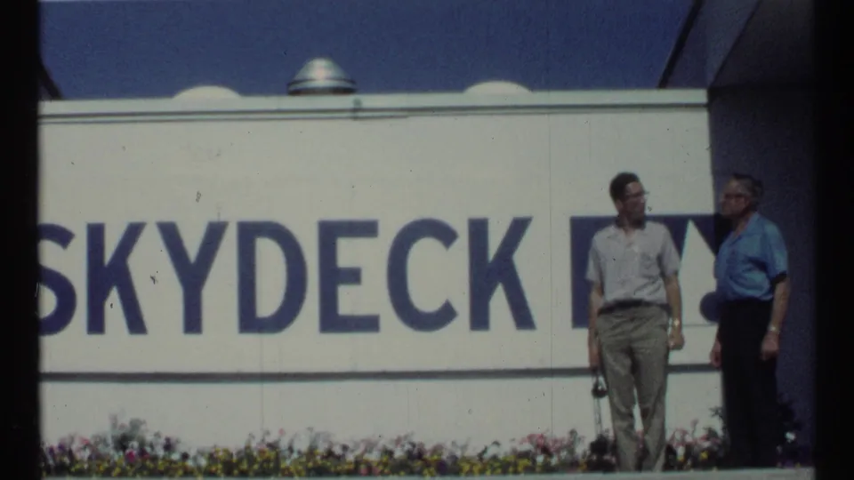 The final frame shows two adult men near a cropped Skydeck sign, with flowers and sunlit white wall panels visible.