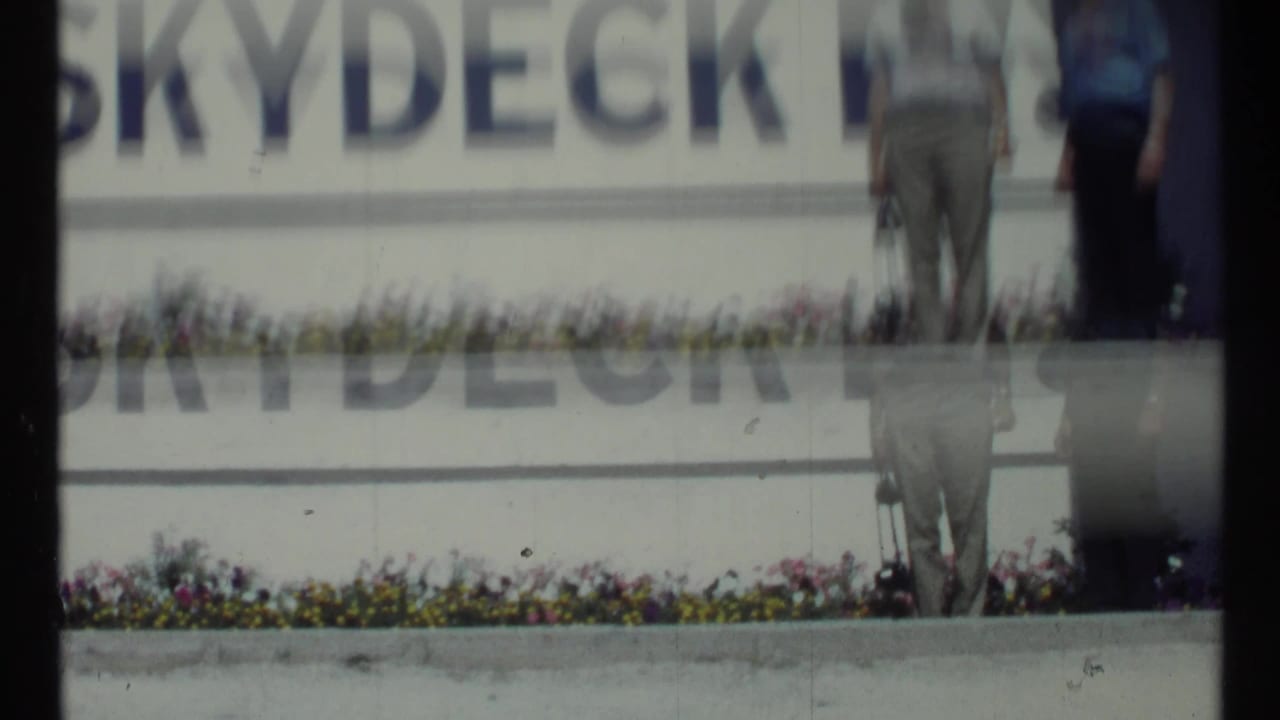 Soft 8mm frame with partial Skydeck sign, flower bed, and blurred standing figures.