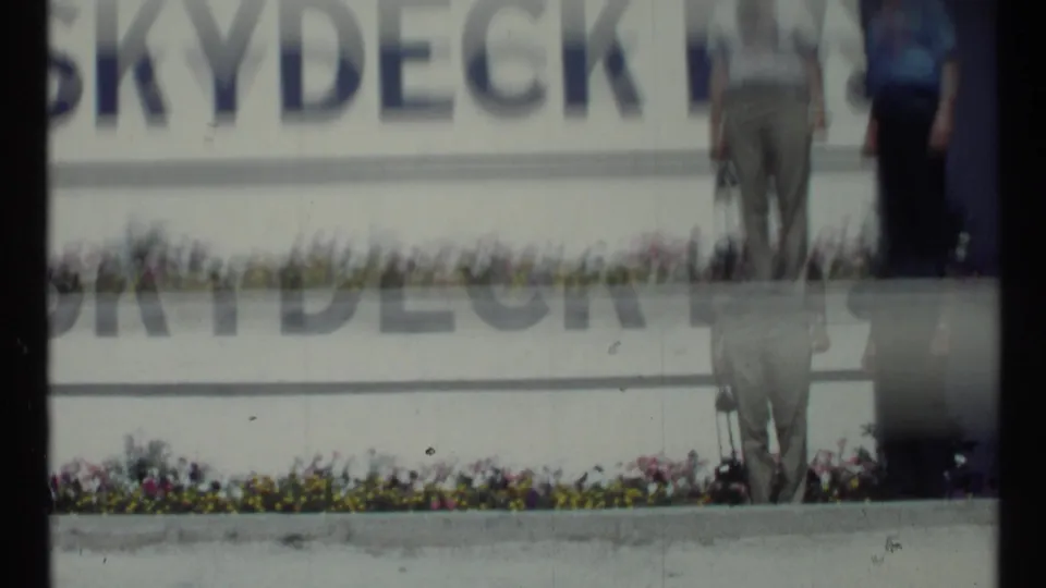 1967 Toronto home-movie frame showing a cropped Skydeck sign with blurred adult figures near a flower bed.