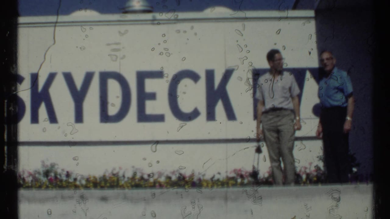 Two adult men stand beside a large Skydeck sign above a low flower bed.
