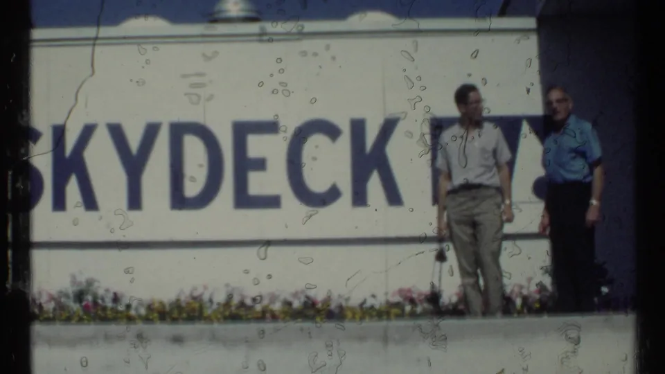 1967 Toronto home-movie frame with two adult men standing by large blue Skydeck lettering on a white wall.