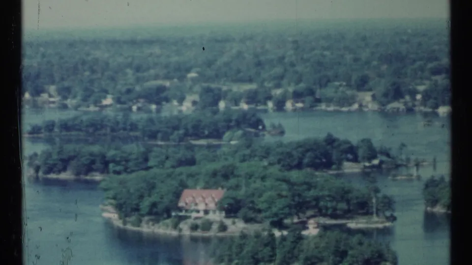 1967 Toronto-area home-movie view looking down toward lake water, trees, shoreline homes, and a red-roofed building.