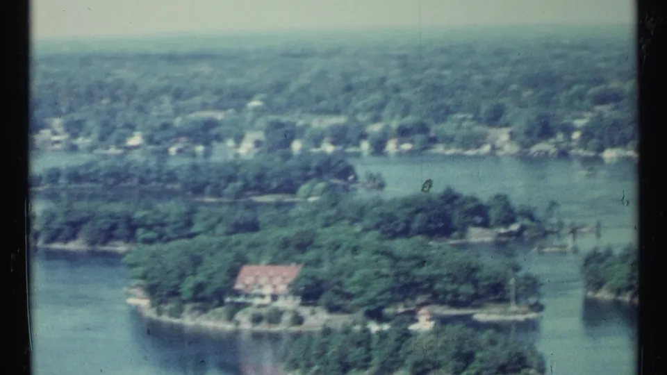 1967 Toronto-area home-movie frame showing an elevated lake view with forest, shoreline houses, and a red-roofed building.