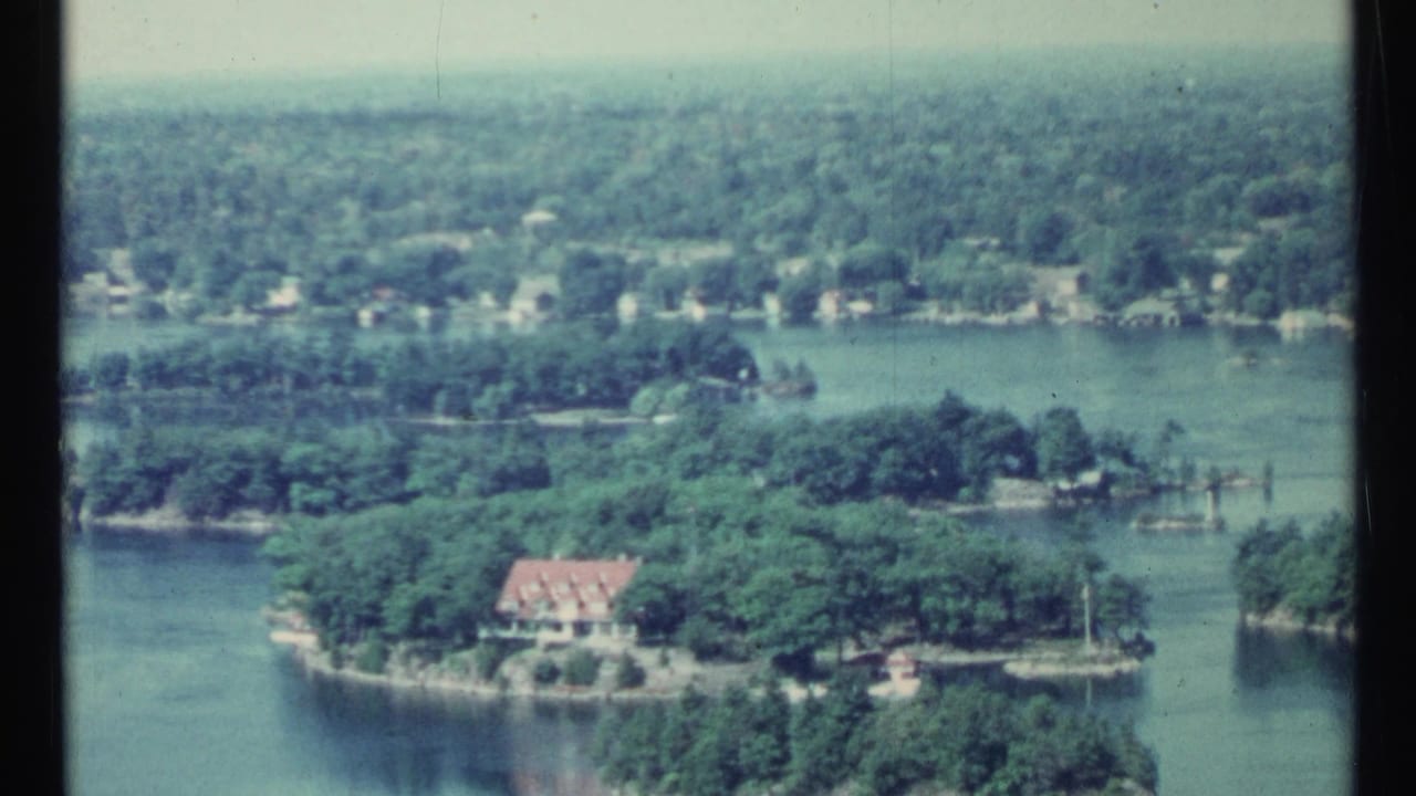 1967 Toronto-area aerial home-movie frame showing wooded islands, calm water, docks, and a red-roofed house on the shoreline.