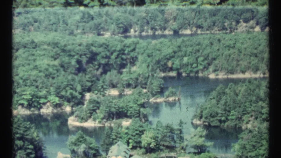 1967 Toronto-area aerial frame showing calm channels, wooded islands, and small shoreline structures in faded 8mm color.