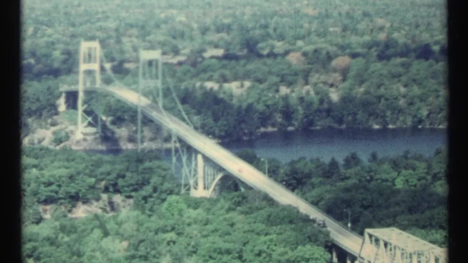 1967 8mm frame showing a suspension bridge over a river, surrounded by dense green vegetation.