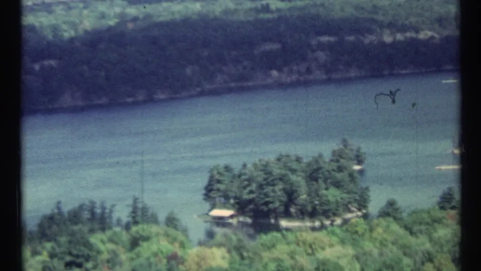 High-angle 8mm view shifts to the river below, showing a wooded shoreline, small structure, and distant opposite bank.