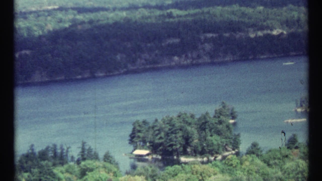 1967 8mm view near Toronto of a calm river, wooded banks, and a small island with a pale roofed structure at the shoreline.