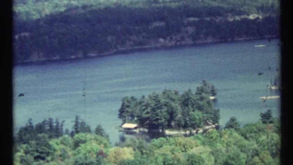 1967 8mm scenic frame near Toronto showing blue-green water, a wooded island, distant shore, and handheld home-movie texture.