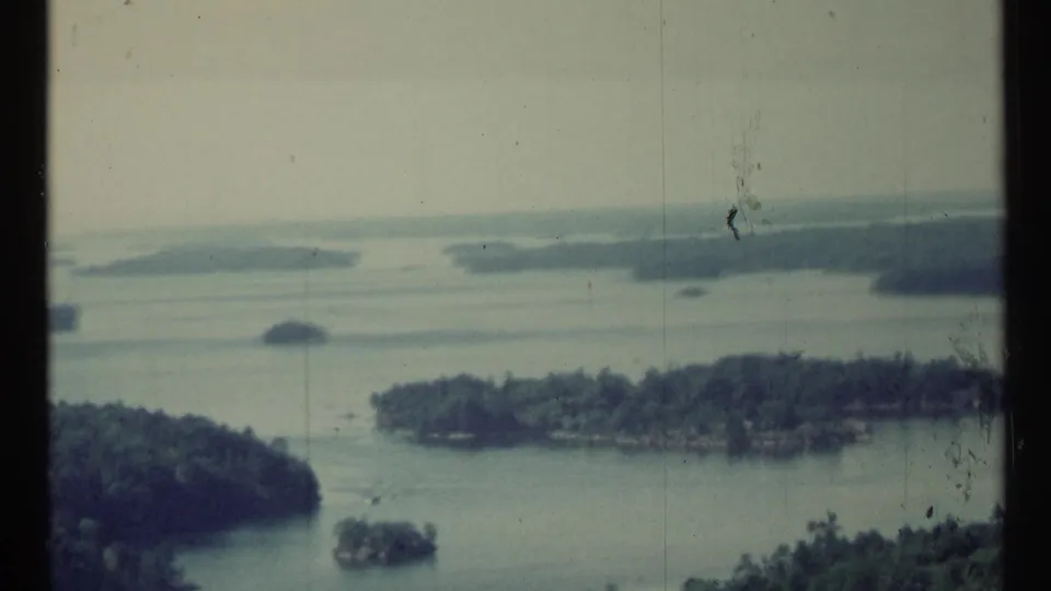 Muted 1967 8mm aerial view of forested islands and lake channels, with soft focus, film dust, and vertical frame wear.