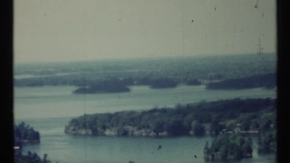 High 1967 8mm view across lake water, wooded shorelines, and small islands, with muted color and visible archival film marks.