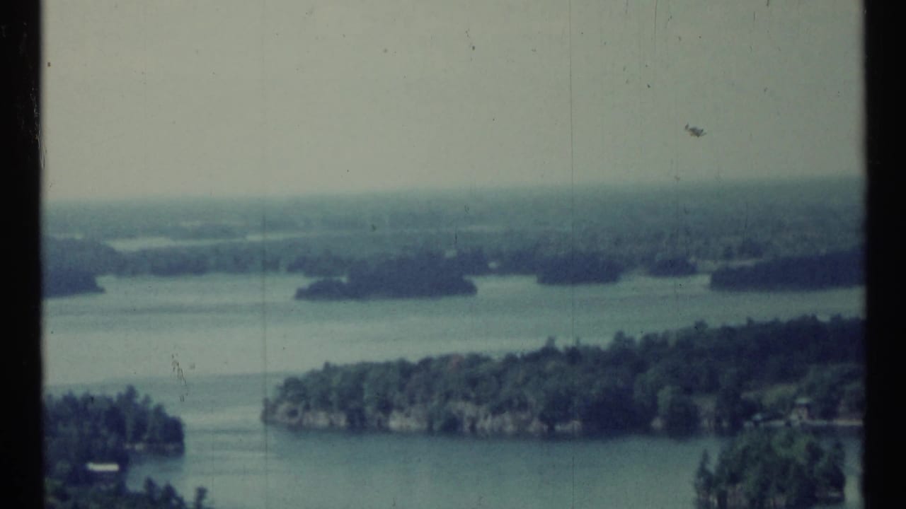 1967 Canadian 8mm frame showing blue-green water, wooded islands, pale sky, and a soft hazy horizon.