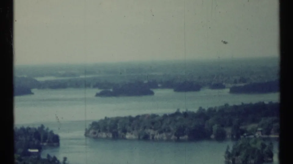 1967 Canadian 8mm frame showing blue-green water, wooded islands, pale sky, and a soft hazy horizon.
