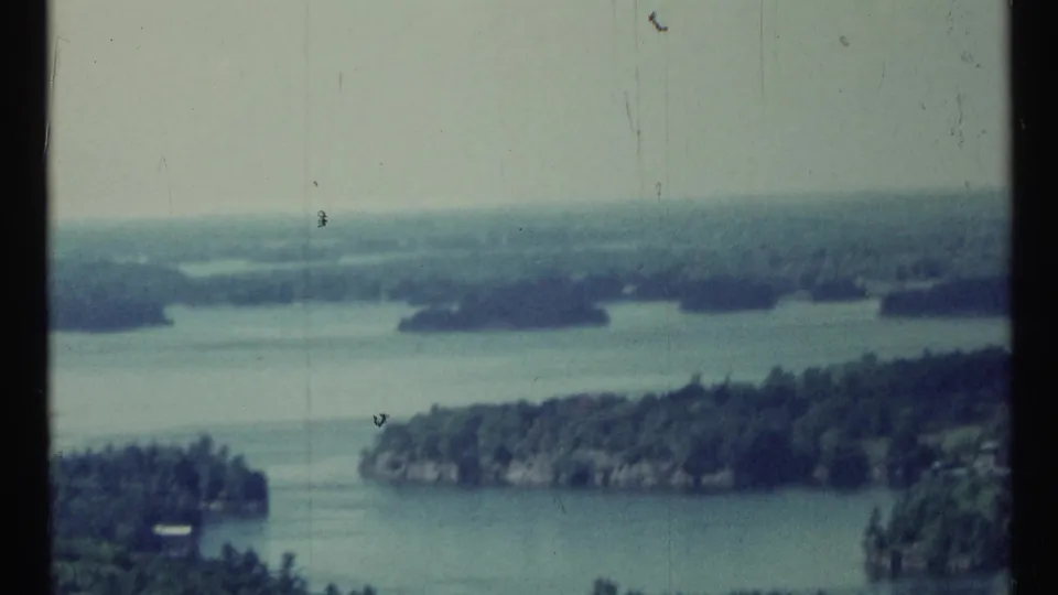 1967 Canadian 8mm frame with a high view across calm water, forested landforms, distant haze, and visible film wear.