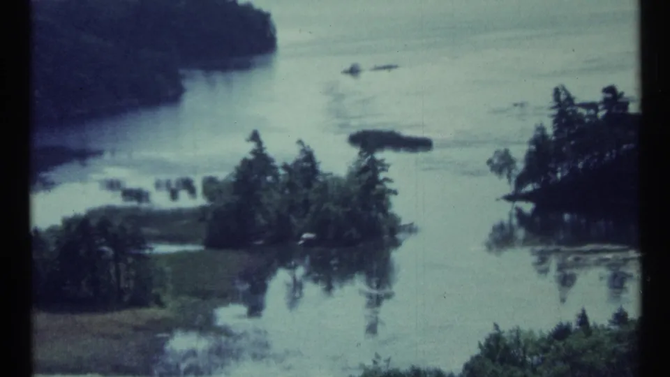 1967 Canadian 8mm frame showing small wooded islands and peninsulas in calm blue-green water, with soft focus and film grain.