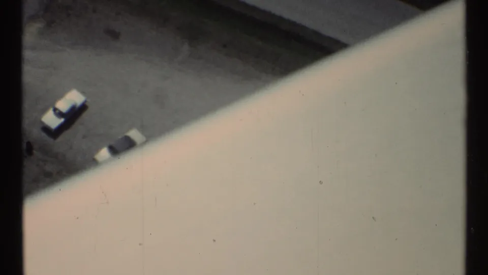 1967 Toronto footage frames two parked cars on asphalt below a broad pale structural edge, with home-movie grain and slight color fading.