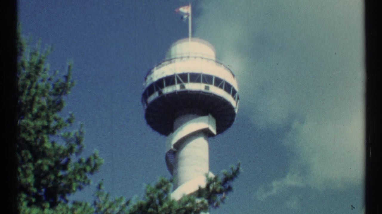 1967 8mm frame of a white tower with a circular observation deck, flags, trees, and clouds in the supplied Toronto metadata.