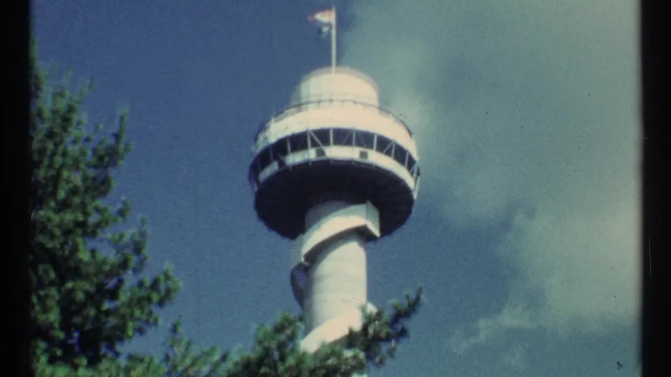 1967 8mm frame of a white tower with a circular observation deck, flags, trees, and clouds in the supplied Toronto metadata.