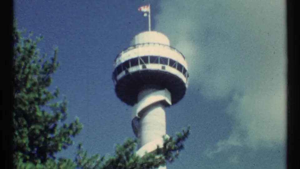 Visitor-shot 8mm view of a white observation tower with a round deck, rooftop flags, tree branches, and blue sky.
