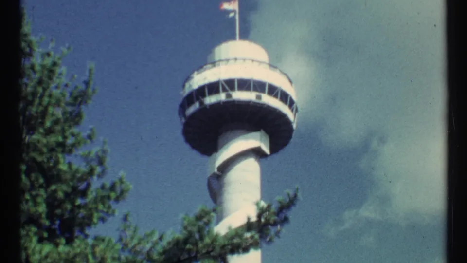 Low-angle 8mm frame showing the white tower, circular deck, flagpole, green branches, and partly cloudy sky.