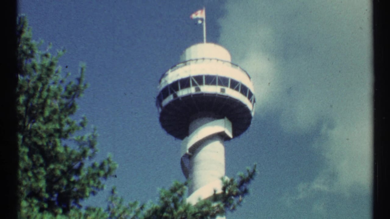 1960s 8mm travel frame showing a circular observation tower with a flagpole, partly obscured by green trees against a blue sky.