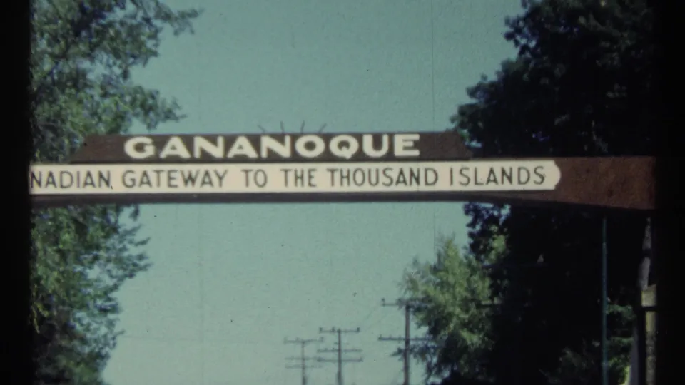 Close 8mm frame of the Gananoque gateway arch, with the Thousand Islands wording partially cropped and trees around the roadside sign.